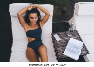 Happy travel blogger in swimwear lying at comfortable sunbed during recreation summer vacations at coastline beach with lounge zone, digital cellular gadget with copy space for your text advertising