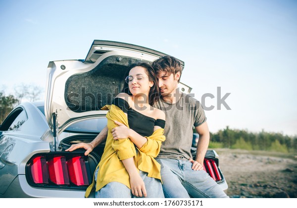 Happy traveler couple sitting in car open trunk
and watch the sunrise