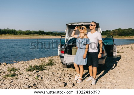 Happy traveler couple standing near car open trunk and watch the sunrise