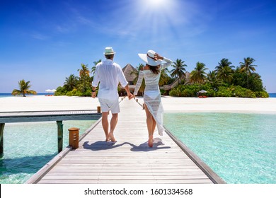 A happy traveler couple in white summer clothing walks down hand in hand a wooden pier towards a tropical paradise island in the Maldives, Indian Ocean - Powered by Shutterstock