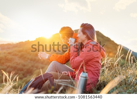 Happy traveler young couple resting in the mountains and drinking tea from thermos at sunset in spring or summer season.