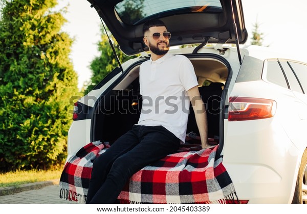 Happy\
traveler, young man with beard and sunglasses, sits in the car,\
open the trunk and watch the sunrise,\
outdoors