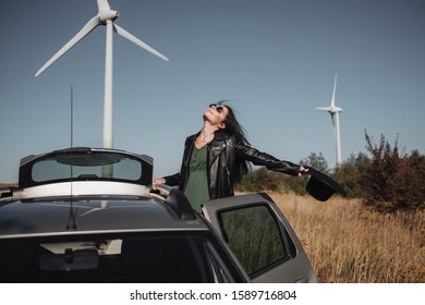 Happy Traveling Girl Enjoying A Car Trip On The Field Road With Electric Wind Turbine Power Generator On The Background