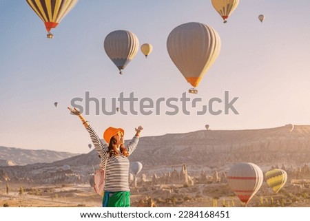 Happy traveller girl raising hands while watching magnificent view of flying hot air balloons in famous tourist attraction - Cappadocia