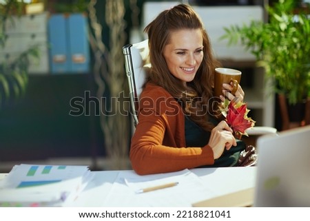 happy trendy small business owner woman with autumn yellow leaves and cup of hot cocoa having video call in the modern green office.