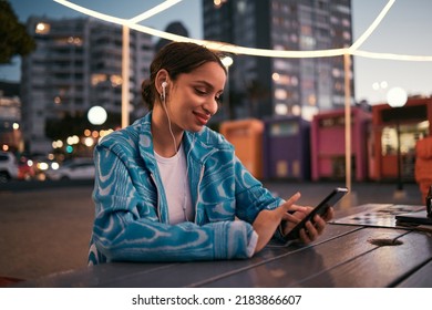 Happy and trendy woman browsing on phone while wearing earphones and listening to music while sitting at outdoor cafe in night city. Happy woman streaming subscription app or making video call - Shutterstock ID 2183866607