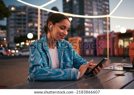 Happy and trendy woman browsing on phone while wearing earphones and listening to music while sitting at outdoor cafe in night city. Happy woman streaming subscription app or making video call
