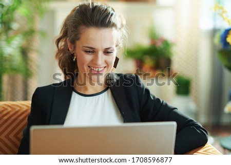 happy trendy woman in white blouse and black jacket in the modern house in sunny day doing research on a laptop while sitting on sofa.