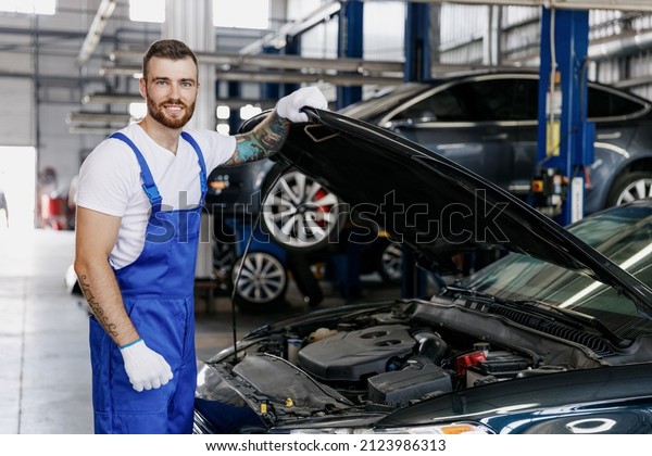 Happy troubleshooter male technician car mechanic man\
in denim blue overalls white t-shirt fix problem with raised hood\
bonnet work in modern vehicle repair shop workshop indoor. Tattoo\
translate fun