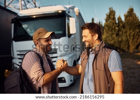 Happy truck driver and his coworker greeting while standing on parking lot. 