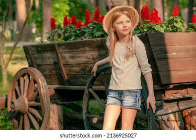 Happy Tween Girl Posing Near Flower Bed In Shape Of Vintage Wooden Cart In Park