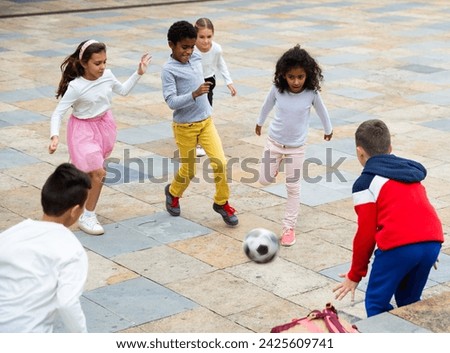 Happy tween girls and boys of different nationalities playing football in schoolyard during break in lessons on warm fall day.