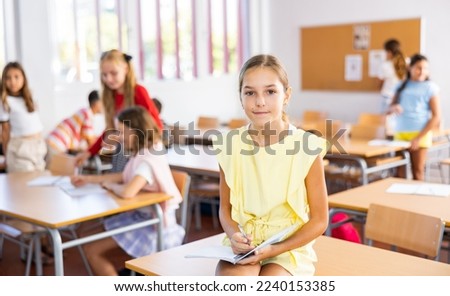 Happy tween schoolgirl repeating educational material before lesson, sitting during break on table with notebook in hands
