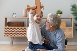 Happy Two 2 Generation Family Old Grandfather And Cute Little Child Boy Grandson Play Hold Wooden Toy Plane Lay On Floor, Funny Small Grandkid Having Fun With Grandpa Fly On Airplane Laughing At Home