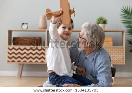 Happy two 2 generation family old grandfather and cute little child boy grandson play hold wooden toy plane lay on floor, funny small grandkid having fun with grandpa fly on airplane laughing at home