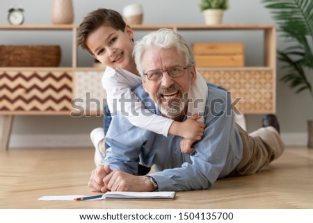 Happy two 2 generations family old grandfather and cute little boy grandson drawing with pencils lying on warm heated wooden floor together, smiling senior grandpa play with grandchild look at camera