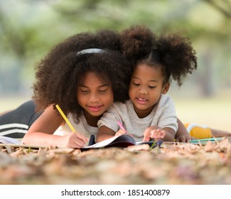 Happy Two African American Little Kids Girl Drawing On Notebook With Pencil While Lying On Mat Outdoor. Two Little Girl Using Pencil Writing And Doing Homework On Notebook Together In The Park