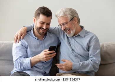 Happy two age generations men family old father embracing young grown adult son having fun enjoying using smart phone bonding watching funny social media video using mobile apps at home sit on sofa