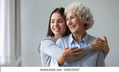 Happy two age generations women family young adult woman daughter granddaughter embrace smiling old elder lady grandma mom look through window intro good future dream enjoy wellbeing together concept