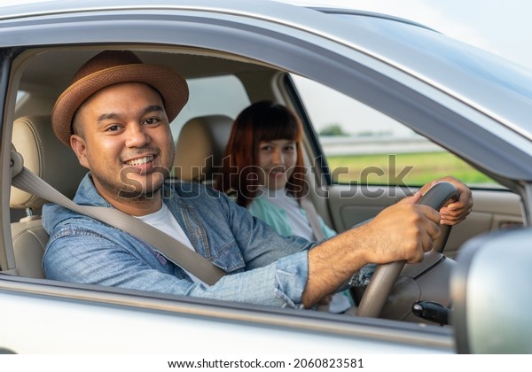 Happy two young friends driver smiling while\
sitting in a car with open front window. Asian man and woman smile\
and looking through window. Young couple driving car to travel on\
holiday vacation time.