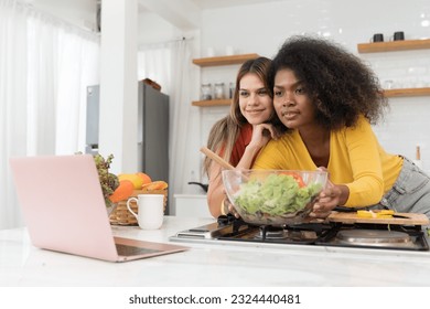 Happy two young women looking laptop computer during cooking together in kitchen room at home. Two young diverse lesbian women spending time together. Diversity, LGBT and gender identity concept - Powered by Shutterstock