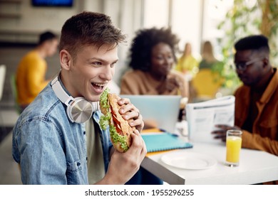 Happy University Student Eating Sandwich Wile Having Lunch Break With Is Friends In Cafeteria.