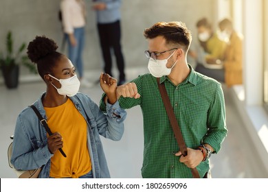 Happy university students greeting in a lobby and elbow bumping while wearing protective face masks. 