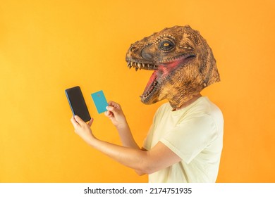 Happy Unrecognizable Person Wearing Dinosaur Animal Head Mask , Holding Smartphone And Credit Card On Orange Background