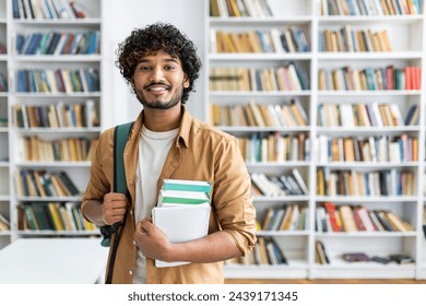 Happy unshaved man wearing backpack and holding stack of books while smiling at camera at bookstore. Optimistic classman making purchases for beginning of new academic semester in college.