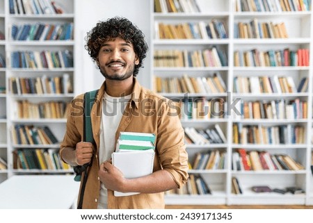 Happy unshaved man wearing backpack and holding stack of books while smiling at camera at bookstore. Optimistic classman making purchases for beginning of new academic semester in college.