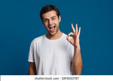 Happy unshaven guy smiling and showing ok sign at camera isolated over blue background