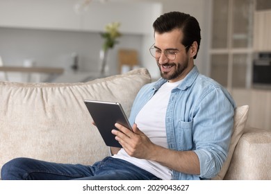 Happy User Holding Digital Device, Resting On Couch, Relaxing At Home. Relaxed Millennial Man Using Tablet Computer, Making Video Call, Reading Book Online, Watching Virtual Content, Smiling At Screen