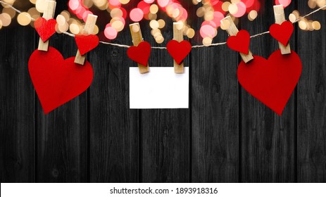 Happy Valentine's Day Background - Red Hearts And White Paper Note Hang On Wooden Clothes Pegs With Wooden Heart And Bokeh Lights On A String Isolated On Black Wooden Wall, With Space For Text