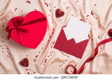 Happy Valentines Day Composition. Flat Lay Red Envelope With Blank Paper Inside, Heart Shaped Box Wrapped Ribbon, Hearts On Pastel Beige Silk Cloth.