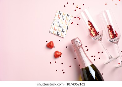 Happy Valentines Day Concept. Romantic Dinner With Champagne Bottle, Glasses, Greeting Card, Heart-shaped Chocolates And Red Confetti On Pink Table. Flat Lay, View From Above.