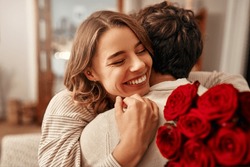 Happy Valentine's Day. A Man Gives A Bouquet Of Red Roses To His Beloved Woman In The Living Room At Home, The Woman Hugging Him Tenderly. Romantic Evening Together.