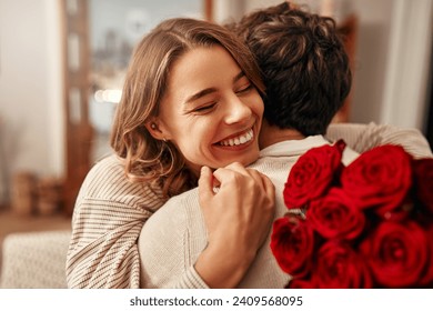 Happy Valentine's Day. A man gives a bouquet of red roses to his beloved woman in the living room at home, the woman hugging him tenderly. Romantic evening together.