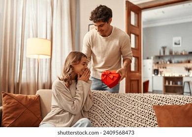 Feliz Día de San Valentín. Un hombre entrega una caja de regalo en forma de corazón a su amada mujer en la sala de estar en casa. Velada romántica juntos.