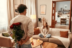 Happy Valentine's Day. A Man Holding A Bouquet Of Red Roses Behind His Back For His Beloved Woman Who Is Sitting On The Sofa In The Living Room At Home.