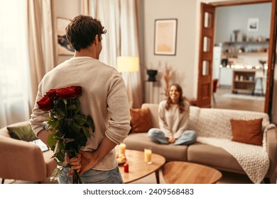 Happy Valentine's Day. A man holding a bouquet of red roses behind his back for his beloved woman who is sitting on the sofa in the living room at home.