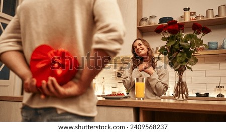 Happy Valentine's Day. A man holding a heart-shaped red gift box behind his back for his beloved woman while standing in the kitchen at home.