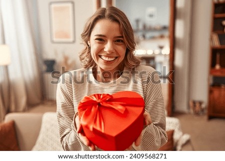 Happy Valentine's Day. Happy smiling woman holding red heart shaped gift box with bow in living room at home.