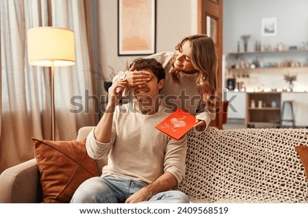 Photo of Happy Valentine's Day. A woman gives a greeting card to her beloved man with the inscription I love you in the living room of the house, covering his eyes with her palm.