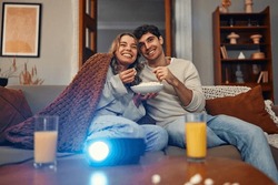 Happy Valentine's Day. A Young Couple In Love Sitting On The Sofa In The Living Room At Home, Eating Popcorn, Covered With A Blanket And Watching A Movie On The Projector.