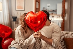 Happy Valentine's Day. Young Couple In Love Holding A Heart-shaped Balloon, Covering Themselves With It While Kissing, Sitting On The Sofa In The Living Room At Home. Romantic Evening Together.