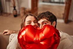 Happy Valentine's Day. Young Couple In Love Holding A Heart-shaped Balloon, Hiding Behind It While Sitting On The Sofa In The Living Room At Home. Romantic Evening Together.