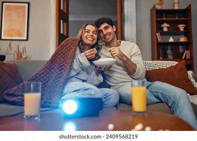 Happy Valentine's Day. A young couple in love sitting on the sofa in the living room at home, eating popcorn, covered with a blanket and watching a movie on the projector. - Powered by Shutterstock