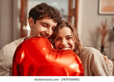 Happy Valentine's Day. Young couple in love holding a heart-shaped balloon, hiding behind it while sitting on the sofa in the living room at home. Romantic evening together.