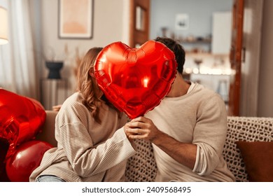 Happy Valentine's Day. Young couple in love holding a heart-shaped balloon, covering themselves with it while kissing, sitting on the sofa in the living room at home. Romantic evening together.