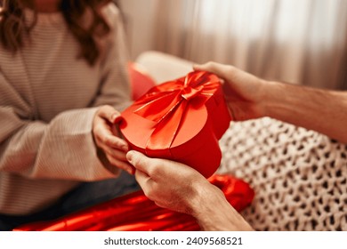 Happy Valentine's Day. Young couple in love exchanging gifts while sitting on the sofa in the living room at home. A man giving a heart-shaped gift box to his beloved woman.